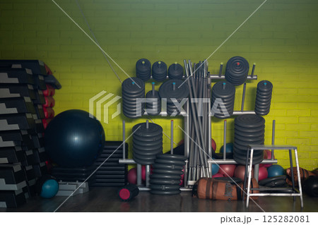 A neatly organized gym equipment storage area featuring a variety of weights, barbells, kettlebells, and fitness accessories against a yellow brick wall. A neatly organized gym equipment storage area featuring a variety of weights, barbells, kettlebells, and fitness accessories against a yellow brick wall. 125282081