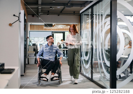 Business colleagues, collaborative business colleagues, including a person in a wheelchair, walk past a modern glass office corridor, illustrating diversity, teamwork and empowerment in the workplace. Business colleagues, collaborative business colleagues, including a person in a wheelchair, walk past a modern glass office corridor, illustrating diversity, teamwork and empowerment in the workplace. 125282188