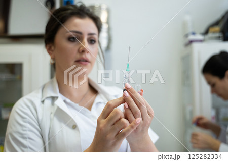 A focused nurse in a white lab coat carefully prepares a syringe for an injection in a professional healthcare environment. A focused nurse in a white lab coat carefully prepares a syringe for an injection in a professional healthcare environment. 125282334