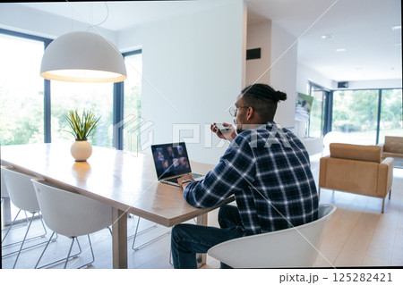 African American businessman in glasses sitting at a table in a modern living room, using a laptop and smartphone for business video chat, conversation with friends and entertainment. 125282421