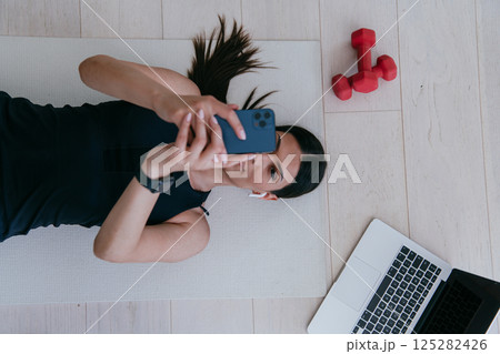 Sportswoman using a smartphone while resting and lying on the floor of modern house. Sportswoman using a smartphone while resting and lying on the floor of modern house. 125282426