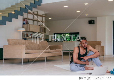 Training At Home. Sporty African American man doing the training while watching an online tutorial on a laptop, exercising in the living room, free space Training At Home. Sporty African American man doing the training while watching an online tutorial on a laptop, exercising in the living room, free space 125282827