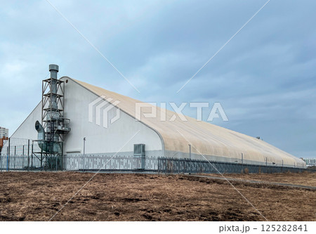 Large industrial building hangar with metal framework and chimney, set against a cloudy sky. The structure includes ventilation systems and is surrounded by sturdy fence on dry land Large industrial building hangar with metal framework and chimney, set against a cloudy sky. The structure includes ventilation systems and is surrounded by sturdy fence on dry land 125282841