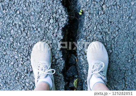 View of two feet in white sneakers standing at the edge of a deep crack in pavement. The scene highlights the condition of worn infrastructure and potential hazards 125282851