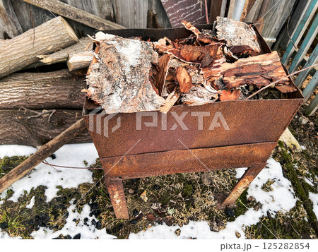 Rusted metal fire pit filled with birch logs and wood pieces. Surrounded by logs and patches of snow, the scene highlights a rustic outdoor setting, perfect for backyard fires and winter warmth 125282854