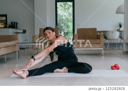 A young woman in sportswear stretching her legs during online training in the living room of her home. A young woman in sportswear stretching her legs during online training in the living room of her home. 125282928