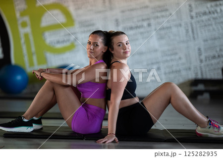 Two fitness women resting and recovering in the gym after an intense workout session, showcasing strength, endurance, and dedication to their fitness journey. Two fitness women resting and recovering in the gym after an intense workout session, showcasing strength, endurance, and dedication to their fitness journey. 125282935