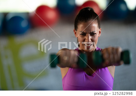 A focused and determined woman performs a strength building dumbbell workout at the gym, showcasing her athletic physique and dedication to fitness. 125282998