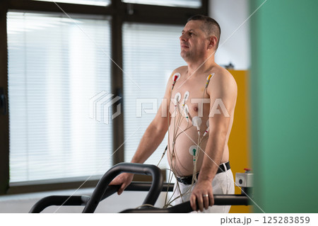 A middle-aged man is undergoing a medical stress test on a treadmill, connected to ECG electrodes for heart monitoring in a clinical environment A middle-aged man is undergoing a medical stress test on a treadmill, connected to ECG electrodes for heart monitoring in a clinical environment 125283859