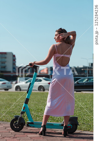 An elegant, stunning woman in a pink dress and sunglasses strikes a confident pose next to her electric scooter, radiating style and sophistication in the vibrant urban environment. 125284236