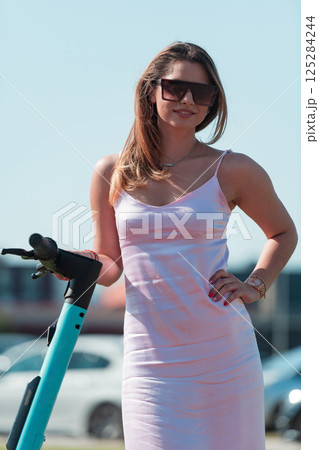 An elegant, stunning woman in a pink dress and sunglasses strikes a confident pose next to her electric scooter, radiating style and sophistication in the vibrant urban environment. An elegant, stunning woman in a pink dress and sunglasses strikes a confident pose next to her electric scooter, radiating style and sophistication in the vibrant urban environment. 125284244