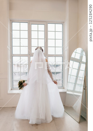 Back view of a Caucasian bride in a white wedding dress and veil standing by a window with a mirror and bouquet Back view of a Caucasian bride in a white wedding dress and veil standing by a window with a mirror and bouquet 125284290