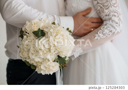 Intimate Wedding Moment: Groom Embracing Bride Holding White Hydrangea Bouquet with Lace Sleeve Details and Elegant Attire, symbolizing love and commitment 125284303