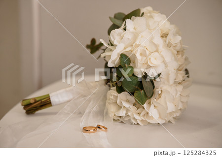 An elegant bridal bouquet composed of lush white hydrangeas and delicate greenery, accompanied by intertwined wedding bands resting on a soft surface symbolizing a commitment 125284325