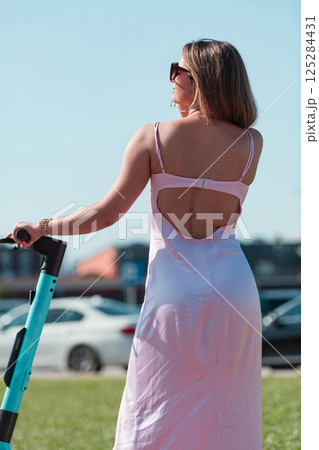 An elegant, stunning woman in a pink dress and sunglasses strikes a confident pose next to her electric scooter, radiating style and sophistication in the vibrant urban environment. An elegant, stunning woman in a pink dress and sunglasses strikes a confident pose next to her electric scooter, radiating style and sophistication in the vibrant urban environment. 125284431