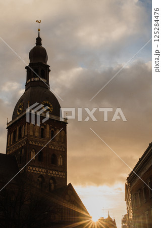 Historic Church Tower with Sunset in the Background.A striking view of a historic church tower silhouetted against a dramatic sky at sunset. The golden sunlight peeks through surrounding buildings 125284476