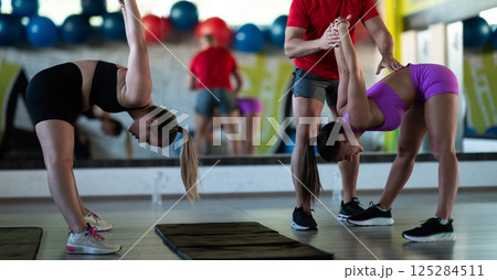 A trainer guides two women through stretching exercises in the gym, promoting flexibility, wellness, and a healthy lifestyle. A trainer guides two women through stretching exercises in the gym, promoting flexibility, wellness, and a healthy lifestyle. 125284511