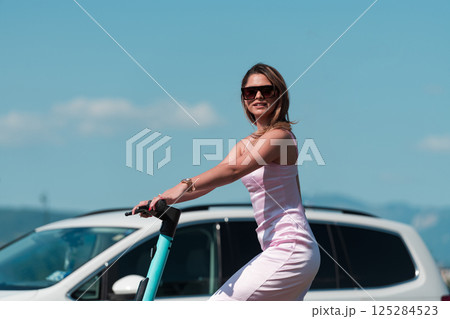 Elegant woman in a pink dress and sunglasses riding an electric scooter through the sunny city streets, embracing the freedom and style of a perfect urban adventure. 125284523