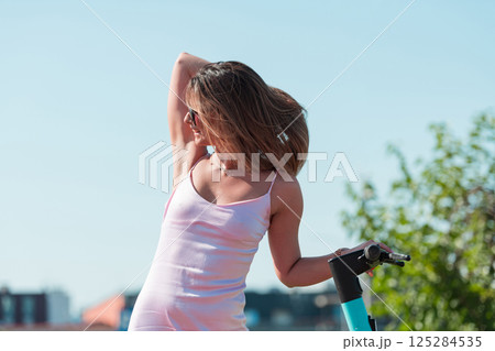 An elegant, stunning woman in a pink dress and sunglasses strikes a confident pose next to her electric scooter, radiating style and sophistication in the vibrant urban environment. An elegant, stunning woman in a pink dress and sunglasses strikes a confident pose next to her electric scooter, radiating style and sophistication in the vibrant urban environment. 125284535