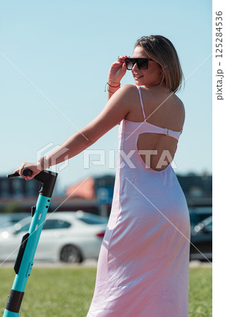 An elegant, stunning woman in a pink dress and sunglasses strikes a confident pose next to her electric scooter, radiating style and sophistication in the vibrant urban environment. 125284536