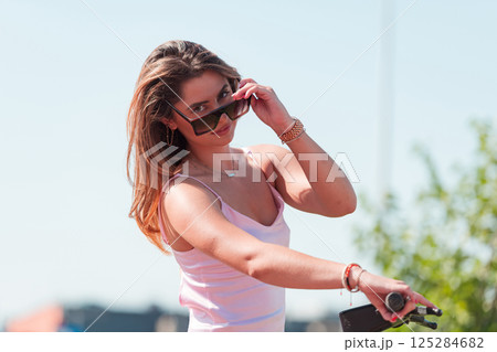 An elegant, stunning woman in a pink dress and sunglasses strikes a confident pose next to her electric scooter, radiating style and sophistication in the vibrant urban environment. An elegant, stunning woman in a pink dress and sunglasses strikes a confident pose next to her electric scooter, radiating style and sophistication in the vibrant urban environment. 125284682