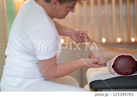 Foot Massage Therapy in a Relaxing Spa Setting. A professional massage therapist in a white uniform provides a soothing foot massage to a client in a calm and serene spa environment 125284703