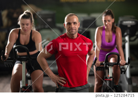 A determined fitness trainer standing confidently in front of his two female clients, guiding them through their workout in the background. 125284715