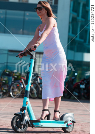 Elegant woman in a pink dress and sunglasses riding an electric scooter through the sunny city streets, embracing the freedom and style of a perfect urban adventure. 125284721