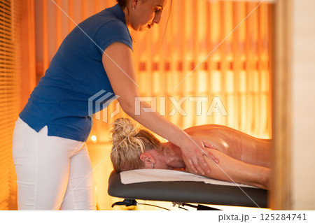 Relaxing Back Massage in a Spa Setting. A professional massage therapist in a blue uniform provides a relaxing back massage to a client lying on a massage table. 125284741