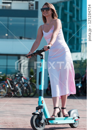 Elegant woman in a pink dress and sunglasses riding an electric scooter through the sunny city streets, embracing the freedom and style of a perfect urban adventure. Elegant woman in a pink dress and sunglasses riding an electric scooter through the sunny city streets, embracing the freedom and style of a perfect urban adventure. 125284754
