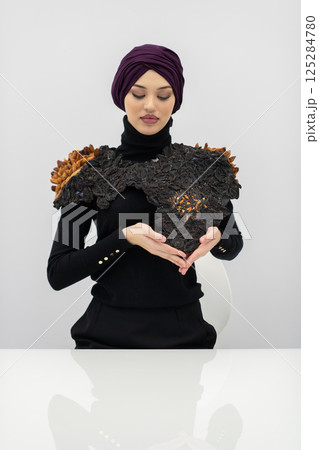 A stylish Muslim woman in a hijab presents her innovative handmade fashion accessory crafted from dried plums in a professional studio. 125284780