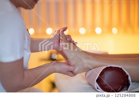 Foot Massage Therapy in a Relaxing Spa Setting. A professional massage therapist in a white uniform provides a soothing foot massage to a client in a calm and serene spa environment Foot Massage Therapy in a Relaxing Spa Setting. A professional massage therapist in a white uniform provides a soothing foot massage to a client in a calm and serene spa environment 125284808
