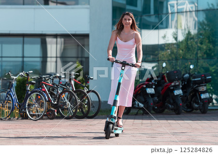 Elegant woman in a pink dress and sunglasses riding an electric scooter through the sunny city streets, embracing the freedom and style of a perfect urban adventure. 125284826