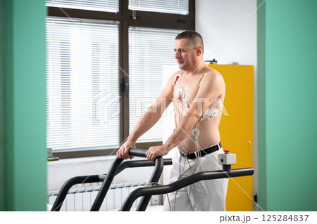 A middle-aged man is undergoing a medical stress test on a treadmill, connected to ECG electrodes for heart monitoring in a clinical environment 125284837