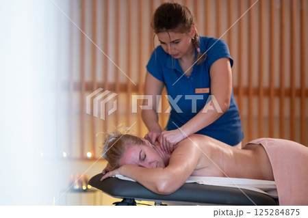 Relaxing Back Massage in a Spa Setting. A professional massage therapist in a blue uniform provides a relaxing back massage to a client lying on a massage table. 125284875