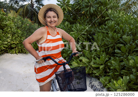 Young woman in casual clothes rides bicycle along path among green bushes. Smiling lady in hat and dress is relaxing among tropical trees. 125286000