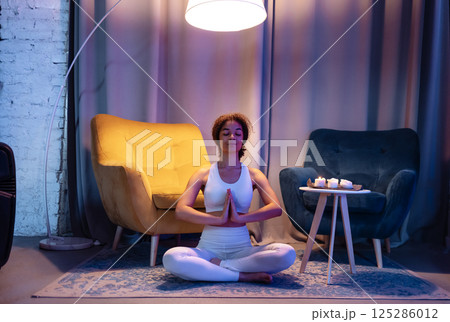 An attractive young African in a white fitness suit sits in a lotus position and relaxes. A cute black girl is meditating in the living room. A relaxed woman does evening yoga at home. An attractive young African in a white fitness suit sits in a lotus position and relaxes. A cute black girl is meditating in the living room. A relaxed woman does evening yoga at home. 125286012