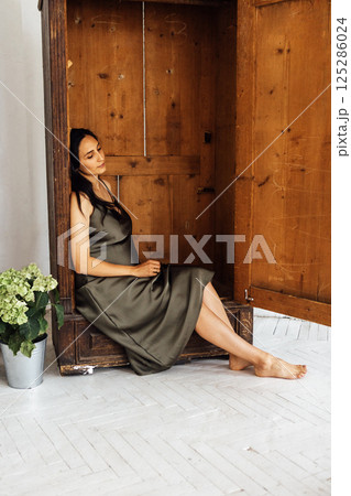 A pregnant woman in a dark green satin dress sits thoughtfully inside a wooden wardrobe. A barefoot lady rests and dreams. A pregnant woman in a dark green satin dress sits thoughtfully inside a wooden wardrobe. A barefoot lady rests and dreams. 125286024