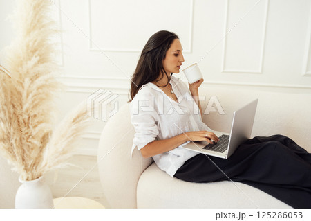 Attractive young businesswoman is sitting on couch and typing on laptop in bedroom. Charming brunette girl works online and drinks coffee at home. Attractive young businesswoman is sitting on couch and typing on laptop in bedroom. Charming brunette girl works online and drinks coffee at home. 125286053