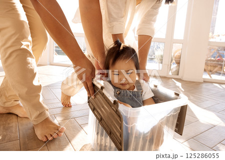 Young laughing Asian parents roll their daughter in a plastic box around the house Young laughing Asian parents roll their daughter in a plastic box around the house 125286055