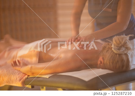 Relaxing Back Massage in a Spa Setting. A professional massage therapist in a blue uniform provides a relaxing back massage to a client lying on a massage table. Relaxing Back Massage in a Spa Setting. A professional massage therapist in a blue uniform provides a relaxing back massage to a client lying on a massage table. 125287210