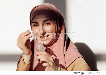 A young girl in a pink hijab applies a transparent moisturizing serum to her face. A teen in a Muslim headscarf smiles and holds a bottle of essence. A young girl in a pink hijab applies a transparent moisturizing serum to her face. A teen in a Muslim headscarf smiles and holds a bottle of essence. 125287300