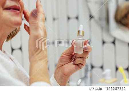 An elderly woman in a white terry dressing gown applies a serum to her face 125287332