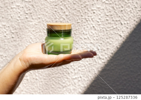 Close-up of a female hand with a jar of green frosted glass moisturizer with a wooden lid. A young woman holds a cosmetic product cream. Close-up of a female hand with a jar of green frosted glass moisturizer with a wooden lid. A young woman holds a cosmetic product cream. 125287336