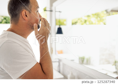 A young man in a white T-shirt puts cream on his cheek in the bathroom. A male looks at himself in the mirror. The man is focused on his skin care routine. A young man in a white T-shirt puts cream on his cheek in the bathroom. A male looks at himself in the mirror. The man is focused on his skin care routine. 125287340