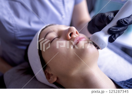 A young woman in a bathrobe lies and receives a facial cleansing procedure using a device A young woman in a bathrobe lies and receives a facial cleansing procedure using a device 125287345