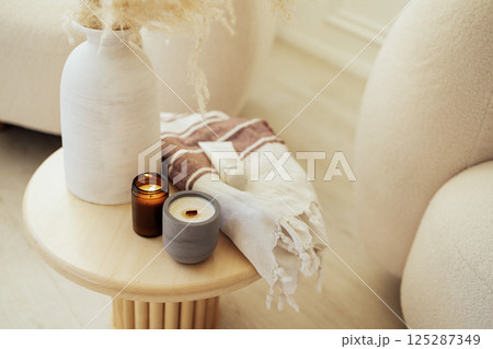 Lit scented candles, mock-up cream, a vase and a blanket on a round coffee table. Cozy home interior in beige tones. 125287349