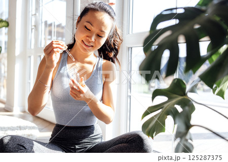 Smiling young Asian woman applies anti-aging serum with vitamins from pipette to her face while sitting in living room. Cute Korean girl takes care of her skin at home. 125287375