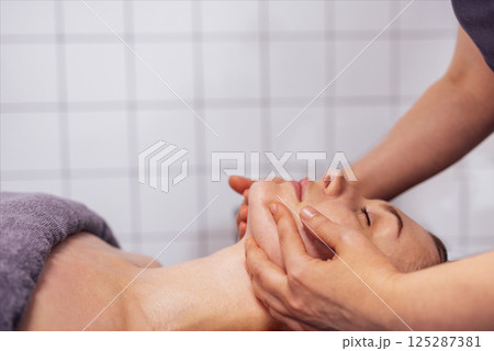 Close up portrait of a young attractive woman getting facial massage. Masseuse hands on the female client chin. Close up portrait of a young attractive woman getting facial massage. Masseuse hands on the female client chin. 125287381