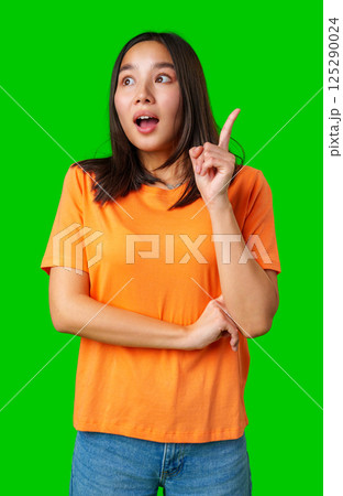 Young woman expressing surprise with one finger raised, wearing an orange shirt against a bright green background 125290024